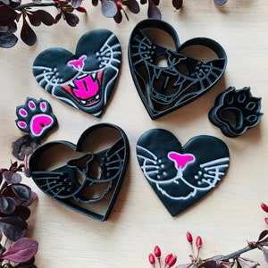 May include: Four black plastic cookie cutters in the shape of a heart with a cat face design. Two cutters have a black outline and white details, and two cutters have a pink nose and mouth. There are also two smaller paw-shaped cutters.