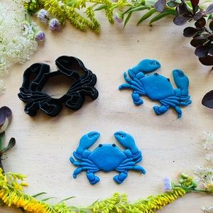 Puede incluir: Dos cortadores de galletas de cangrejo azul y un cortador de galletas de cangrejo negro sobre una superficie de madera rodeada de flores verdes y amarillas.