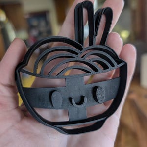 Puede incluir: Un cortador de galletas de plástico negro con forma de cuenco de fideos ramen con una cara sonriente. El cortador tiene una parte superior curvada, una banda rectangular y dos formas curvas en la parte superior, que se asemejan a orejas de conejo.