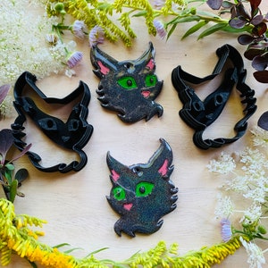 以下が含まれることがあります： 黒いプラスチック製の猫型クッキーカッター2個と、緑色の目とピンクの耳を持つ黒いラメ入りの猫型オーナメント2個。オーナメントの上部には吊り下げ用の小さな穴があり、明るい木製の表面に配置されています。