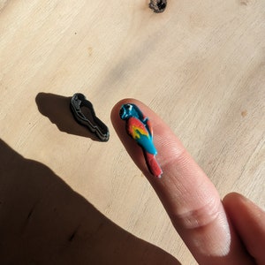 Könnte beinhalten: Ein farbenfroher Papagei-förmiger Polymer Clay Charm, mit blauen, roten, gelben und grünen Farbtönen. Daneben befindet sich ein schwarzer Papagei-förmiger Ausstecher. Der Charm wird auf einem Finger gehalten.