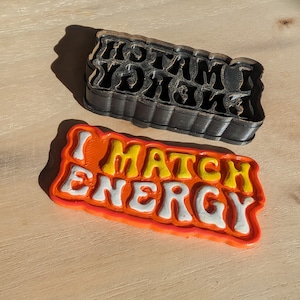 Puede incluir: Un letrero de plástico naranja y amarillo con el texto "I Match Energy" en letras blancas. El letrero está ligeramente curvado y tiene un efecto 3D.