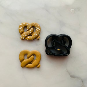 Puede incluir: Dos cortadores de galletas con forma de pretzel, uno dorado y otro negro. El cortador dorado tiene una superficie texturizada y el cortador negro tiene una superficie lisa.