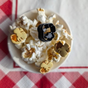 May include: A white bowl filled with popcorn and three small, yellow, cookie-like treats. A black plastic ring is in the centre of the bowl.