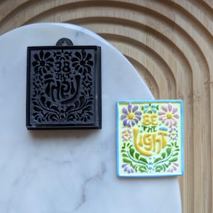 May include: Two square ceramic tiles with floral designs. The tile on the left is black with white lettering that says "38 in the" and the tile on the right is light blue with a yellow background and white lettering that says "Be the Light".