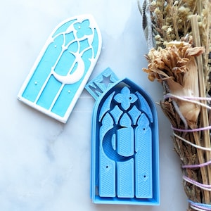 May include: A light blue 3D printed window frame with a crescent moon and star design. The frame is open and the back is visible.