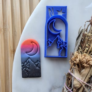 May include: A set of two blue plastic cookie cutters, one with a crescent moon and mountain design, and the other with a sunset and mountain design.