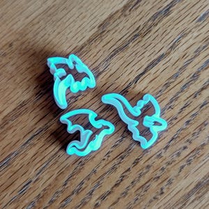May include: Three teal plastic bat-shaped cookie cutters are displayed on a wooden surface. The cutters feature a simple outline design with wings and pointed ears, ideal for Halloween baking. The cutters are in the shape of bats.