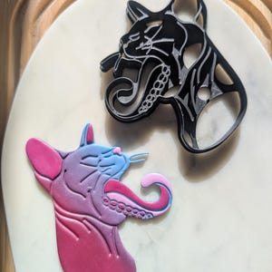 Puede incluir: Dos cortadores de galletas con forma de gato, uno negro y otro rosa, azul y morado. El cortador negro es una silueta detallada de un gato con una lengua larga. El cortador rosa, azul y morado es un gato más caricaturesco con una lengua larga y rizada.
