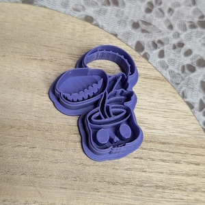 May include: A purple plastic cookie cutter in the shape of a skull with a coffee cup design. The cutter has a handle and is placed on a wooden surface. The skull has eye sockets and a mouth.