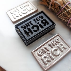 May include: Three rectangular stamps with the text "Eat the Rich" in different styles. The stamps are made of different materials, including black plastic, white plastic, and a metallic gold color.