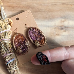 May include: A pair of dangle earrings with a brown, purple, and gold design featuring owls and forest scenes. A small black owl charm is also pictured.