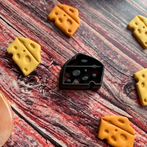 May include: An assortment of cheese-shaped polymer clay pieces and a cheese-shaped cookie cutter. The clay pieces are yellow-orange with holes, resembling Swiss cheese. The cutter is black plastic. The items are arranged on a wooden surface.