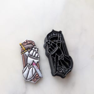 Könnte beinhalten: Zwei Ausstecher, einer ist silberfarben und zeigt eine Hand, die ein Messer mit einem roten Fleck hält, der andere ist schwarz und zeigt eine Hand, die einen Bogen und einen Pfeil hält.