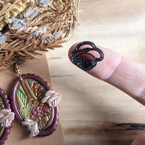 May include: A pair of oval-shaped earrings with a purple and gold iridescent finish. The earrings feature a 3D design of mushrooms and leaves. A small black mushroom-shaped cookie cutter is also shown.