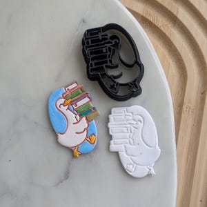 Puede incluir: Tres cortadores de galletas, uno negro, uno blanco y uno de color, todos representando un pato de dibujos animados que lleva una pila de libros. El cortador de color tiene un fondo azul y el pato es blanco con un pico amarillo y patas naranjas.