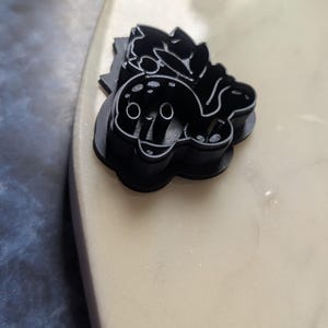 Peut inclure: Emporte-pièce en plastique noir en forme de baleine de dessin animé avec une fleur sur la tête. La baleine a de grands yeux et une bouche souriante. Le découpoir est sur une surface de couleur claire.
