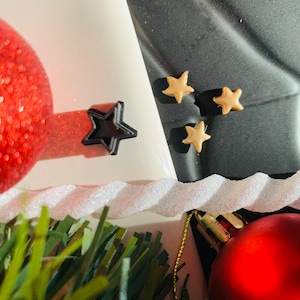 May include: A black star-shaped cookie cutter and three small, light tan star-shaped decorations. The items are arranged near a red glittery ornament, a white twisted decorative element, and green artificial pine needles.