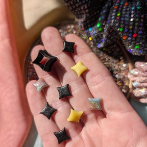Puede incluir: Una colección de pequeños pendientes en forma de diamante brillantes en negro, plata y amarillo.