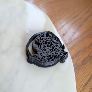 Puede incluir: Cortador de galletas negro de plástico con un intrincado diseño floral y de calavera. El cortador es circular y presenta rosas, hojas y una calavera. Ideal para crear galletas detalladas, el diseño es intrincado y adecuado para hornear.