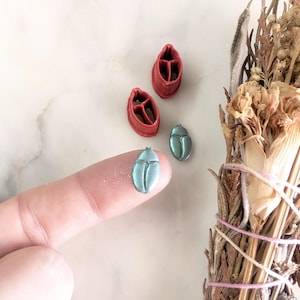 Puede incluir: Dos amuletos de escarabajo verde y dos moldes de plástico rojo para hacer amuletos de escarabajo.