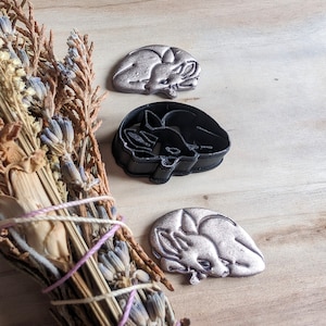 Puede incluir: Tres cortadores de galletas de zorro, uno plateado, uno negro y uno dorado. Los cortadores están sobre una superficie de madera con un manojo de lavanda y salvia secas.