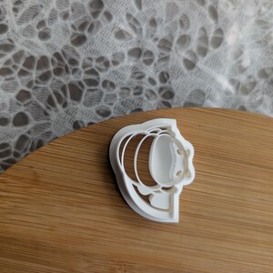 May include: A white plastic cookie cutter featuring a stylized bee design. The cutter has a rounded body shape with horizontal stripes, two small circular eyes, and a pair of wings extending from the sides, creating a clear outline for baking.