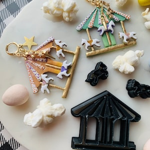 Puede incluir: Dos pendientes de carrusel dorados con caballos coloridos y un dije de estrella. También se muestra un cortador de galletas de carrusel negro.