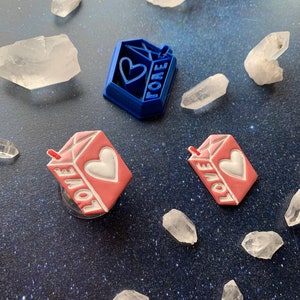 May include: A blue and pink decorative set featuring the word "FORE" and "LOVE". The set includes a blue pendant with a heart and the word "FORE", and two pink milk carton-shaped accessories with the word "LOVE" and a heart.