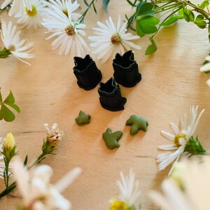May include: Three black cookie cutters shaped like leaves, and four green leaf-shaped cookie cutters on a light brown wooden surface. White flowers surround the cookie cutters.