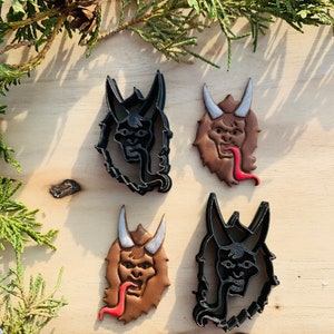May include: Four cookie cutters in the shape of a monster's face with horns and a tongue sticking out. Two of the cutters are black and two are brown.