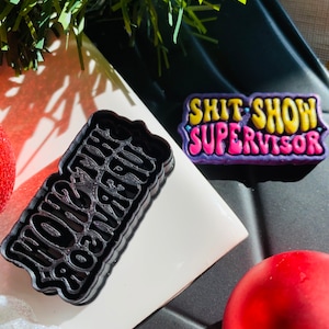 Puede incluir: Cortador de galletas negro y un pin colorido con las palabras "SHIT SHOW SUPERVISOR". El cortador de galletas es negro y el pin tiene colores amarillo, rosa y morado. Los artículos están sobre una baldosa blanca y una superficie negra.