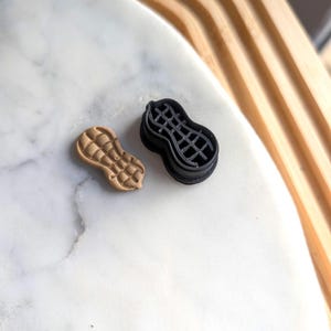 以下が含まれることがあります： ピーナッツの形をしたクッキーカッターが2つ、1つは茶色、もう1つは黒色です。茶色のカッターはソリッドで、黒いカッターはグリッドパターンになっています。
