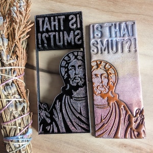 May include: Two black and brown rectangular signs with a glittery finish. The top sign reads "Is That Smut?!" and the bottom sign reads "Is That Smut?!".