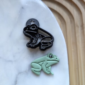 May include: Two frog figurines, one black and one green. The black frog has a star on its head and is made of plastic. The green frog is made of polymer clay.