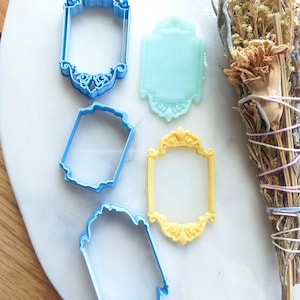 Puede incluir: Cuatro cortadores de galletas de diferentes tamaños en forma de marcos ornamentados. Los cortadores son azules, amarillos y verde claro.