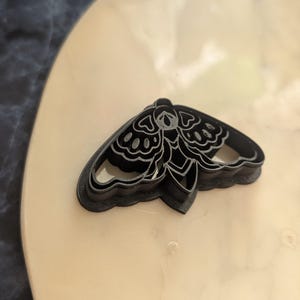 May include: Black plastic moth-shaped cookie cutter. The cutter features detailed wing patterns and a rounded body. The cookie cutter is designed for baking and crafting. The cutter is on a light-colored surface.