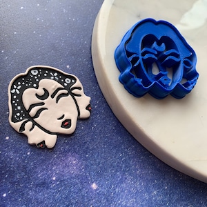 May include: A blue plastic cookie cutter in the shape of a woman's face with a crescent moon on her forehead. The cookie cutter is on a white surface. There is a white cookie with a black and white design on the surface next to the cookie cutter.