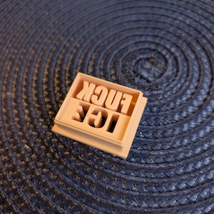 May include: A light peach-colored cookie cutter with the words "ICE FUCK" in capital letters. The letters are raised and the cutter is square-shaped. The cutter is sitting on a dark blue woven surface.