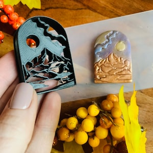 Könnte beinhalten: Eine schwarze Plastikform zum Herstellen von Polymer Clay-Anhängern. Die Form ist wie ein Bogenfenster geformt mit einer Bergszene im Inneren. Die Szene beinhaltet einen Berg, einen Mond und Wolken.