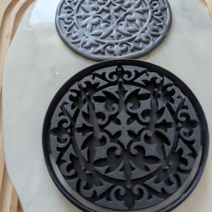 Könnte beinhalten: Zwei schwarze, runde, dekorative Ausstecher mit komplizierten Blumenmustern. Die Ausstecher befinden sich auf einer weißen Marmorfläche.