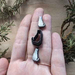May include: Three black and silver cat-shaped cookie cutters. The cutters are in the shape of a cat sitting upright, a cat sitting with its tail curled around its legs, and a cat sitting with its tail straight out.