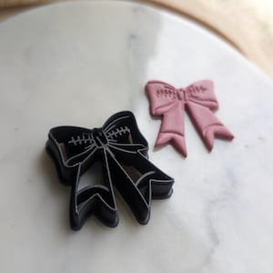 Puede incluir: Dos cortadores de galletas con forma de lazos. El cortador más grande es negro y el más pequeño es rosa.