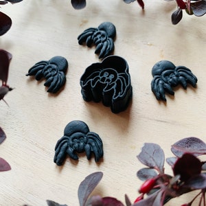 May include: Five black plastic spider cookie cutters on a light brown wooden surface. The cutters are in various sizes and shapes, with some having more detailed features than others. The surface is surrounded by a branch of dark red leaves.