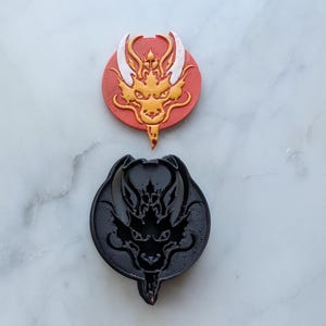 May include: A red and gold dragon cookie cutter with a black dragon cookie cutter next to it. Both cutters are on a white marble surface.