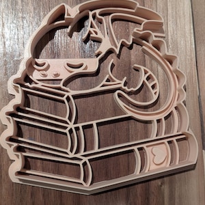 Puede incluir: Cortador de galletas marrón claro con forma de dragón enroscado sobre una pila de libros. Los libros tienen forma de corazón en el lateral. El dragón tiene un diseño detallado con cola curva y alas.