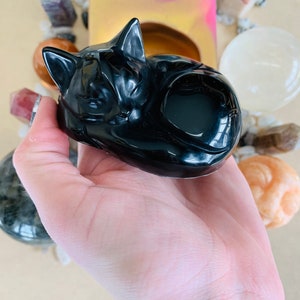 May include: A black obsidian cat figurine, carved in a sleeping pose. The figurine is smooth and polished, with a glossy finish.