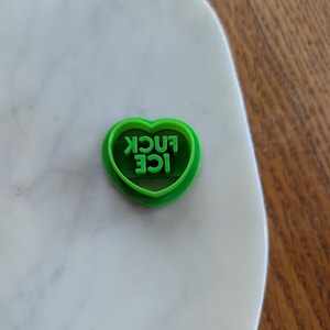 May include: A bright green heart-shaped cookie cutter with the words "ICE FUCK" in capital letters. The cookie cutter is made of plastic and sits on a white marble surface.
