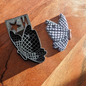 May include: A black butterfly-shaped cookie cutter with a star and the letters "MW" cut out of the top. Next to it is a gray butterfly-shaped object with a textured surface and a small brown piece.