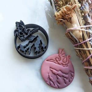 Puede incluir: Un cortador de galletas negro con forma de dragón junto a una galleta con forma de dragón. El cortador es de forma circular con un diseño de dragón y montaña. La galleta es redonda con un diseño de dragón y montaña y es de color rosa.